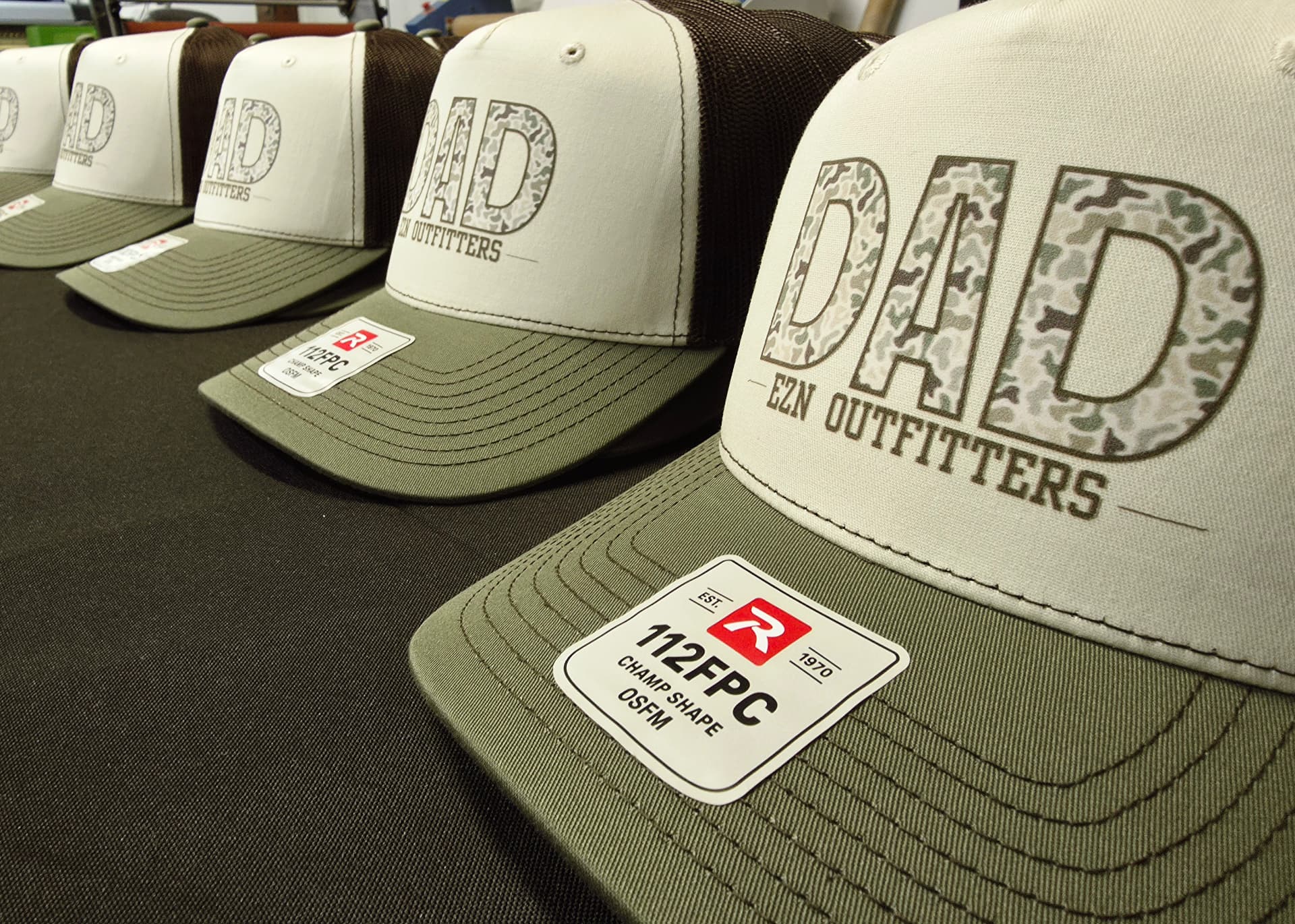 Row of tan and olive trucker hats with camo fill DAD EZN Outfitters print